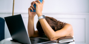 A woman overwhelmed by work, resting head on table with laptop, phone, and smartwatch